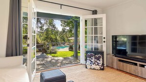 A modern living room with a TV, a sofa, and a glass door leading to a backyard with a pool. - Oak and Stone | Pool + Glen Ellen Wine Country (Glen Ellen)