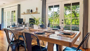 A dining area with a wooden table set for four, chairs, a potted plant, and a view through large windows. - Oak and Stone | Pool + Glen Ellen Wine Country (Glen Ellen)