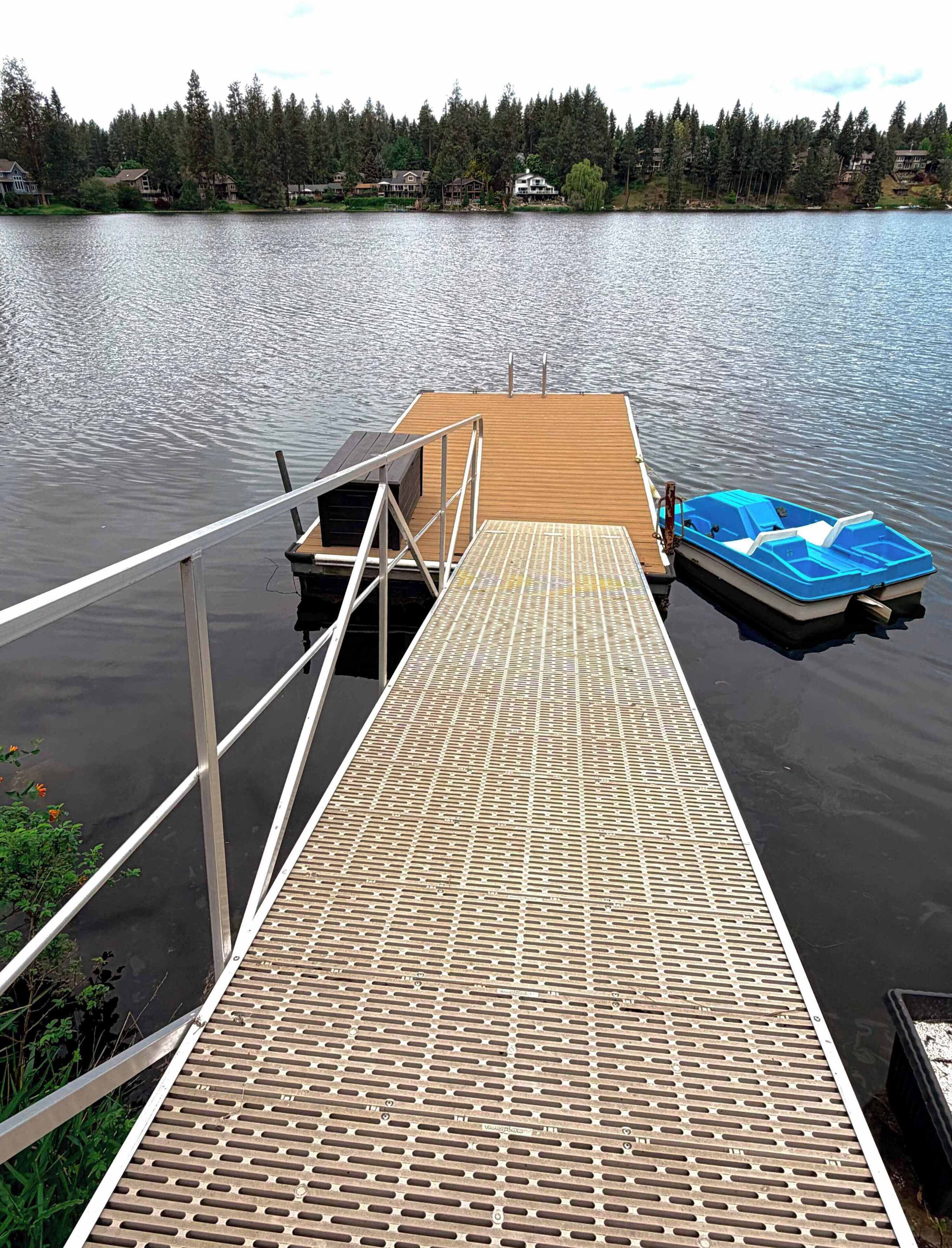Une jetée en bois s’étendant sur une étendue d’eau calme, une petite barque bleue amarrée à son extrémité, et une rive boisée en arrière-plan.