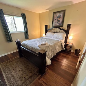 Une chambre Ă coucher avec un lit, une table de chevet, une lampe, une fenĂȘtre avec des rideaux, un tableau au mur et un tapis.