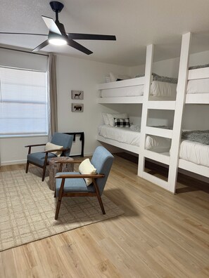 A room with bunk beds, a ceiling fan, a window with blinds, and two armchairs.