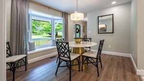 A dining area with a round table, black chairs, a chandelier, and a view of greenery outside.