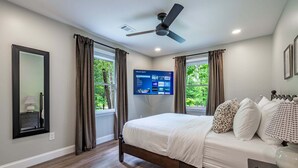 A bedroom with a bed, a ceiling fan, a TV mounted on the wall, and a window with curtains.