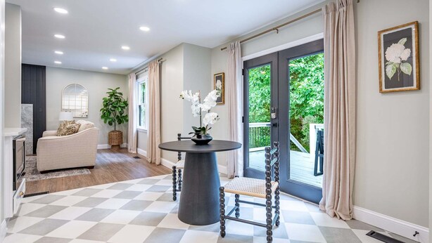 A hallway with a checkered floor, a round table with chairs, a sofa, a potted plant, and a door leading to an outdoor area.