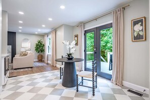 A hallway with a checkered floor, a round table with chairs, a sofa, a potted plant, and a door leading to an outdoor area.