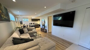Een moderne woonkamer met een ruime hoekbank, een flatscreen televisie aan de wand en een keuken op de achtergrond.