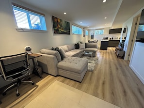 Un salon moderne avec un canapé d’angle, une table basse en verre et un bureau avec une chaise. On aperçoit, au fond, un coin cuisine avec une table et des chaises.