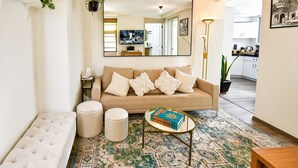 Ein Wohnzimmer mit einem beigen Sofa, einem runden Couchtisch, einem gemusterten Teppich und einem an der Wand montierten Fernseher.