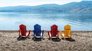 Four Adirondack chairs on a pebbled beach with a lake and mountains in the background.