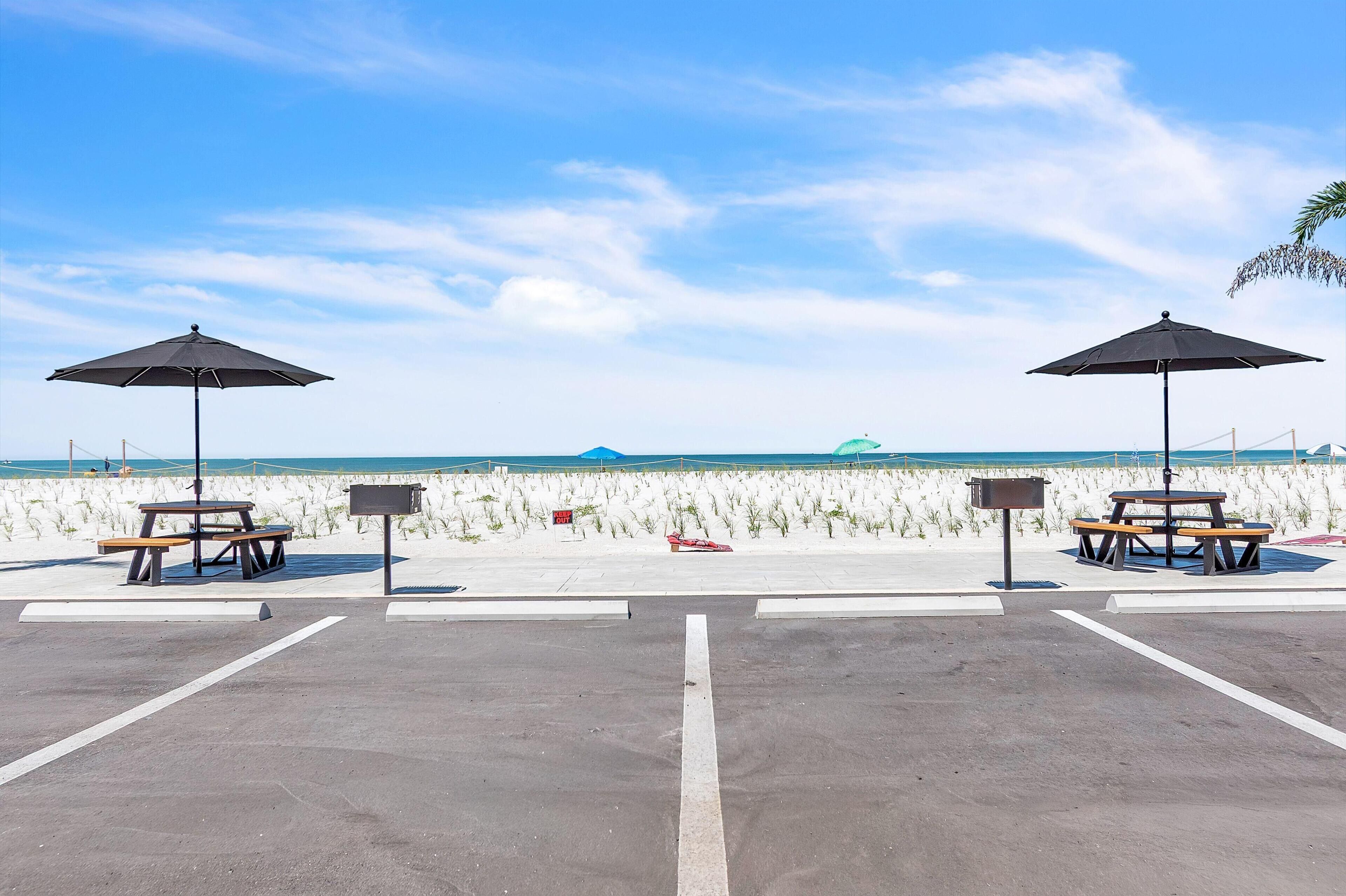 A beachside parking area with picnic tables and umbrellas, a clear sky, and a sandy beach in the background.