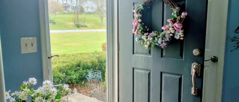 Eine blaue Tür mit einem Kranz, ein Fenster mit Blick auf eine Rasenfläche und Häuser, und eine Vase mit Blumen auf dem Boden.