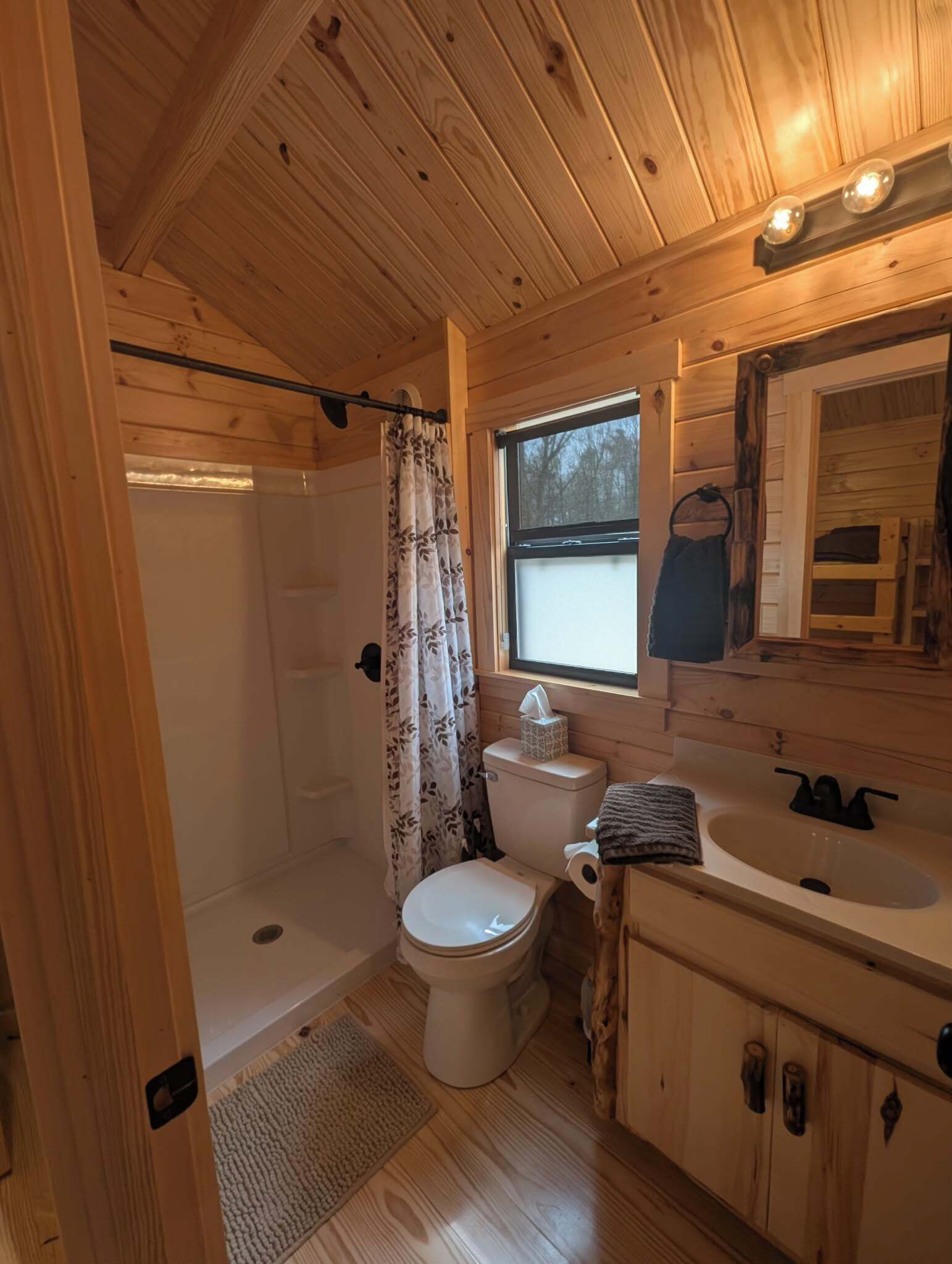 A wooden cabin bathroom with a shower, toilet, sink, and window.