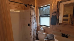 Un baño rústico en una cabaña de madera, con ducha, inodoro y lavamanos, y una ventana.