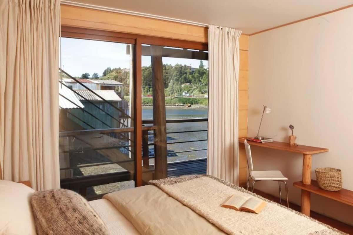 A bedroom with a large window, a bed, a desk, and a chair.