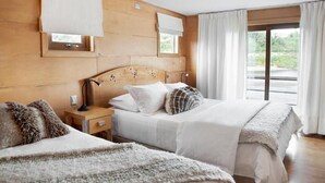 Un dormitorio de hotel con cabecera de madera, una cama grande con ropa blanca, una mesita de noche con lámpara y vistas al verdor por la ventana.