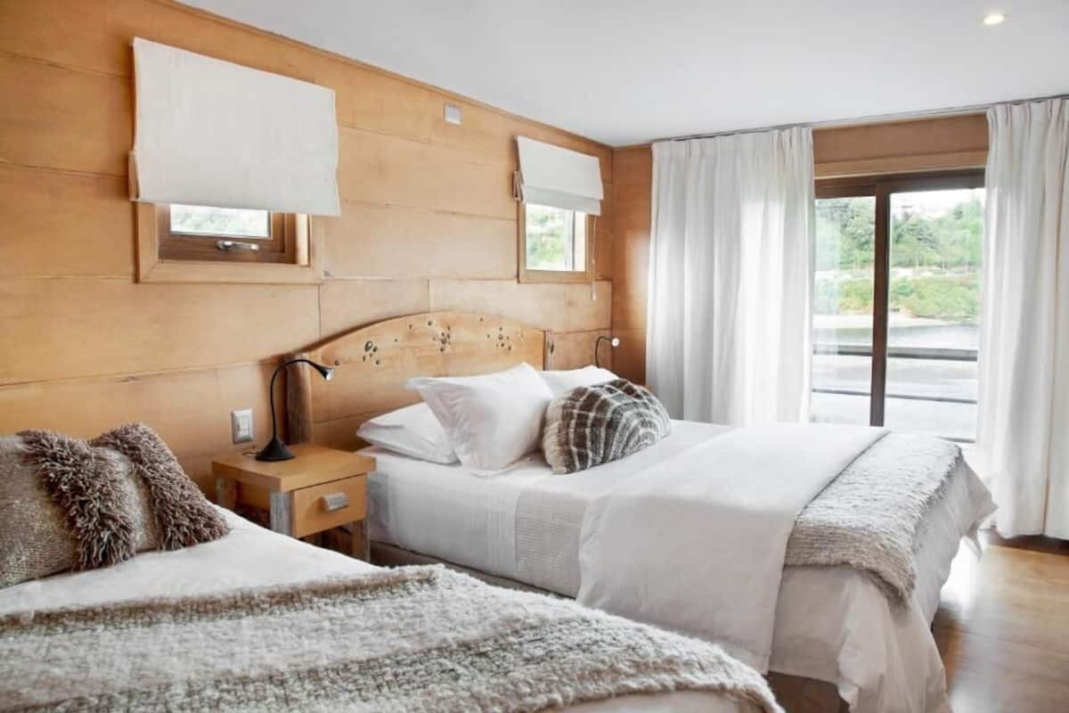 A hotel room with a wooden headboard, a large bed with white bedding, a nightstand with a lamp, and a view of greenery through the window.