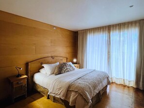 Ein Schlafzimmer mit hölzernem Kopfende, einem Bett mit weißer Bettwäsche, einer Nachttischlampe und einem Fenster mit durchsichtigen Vorhängen.