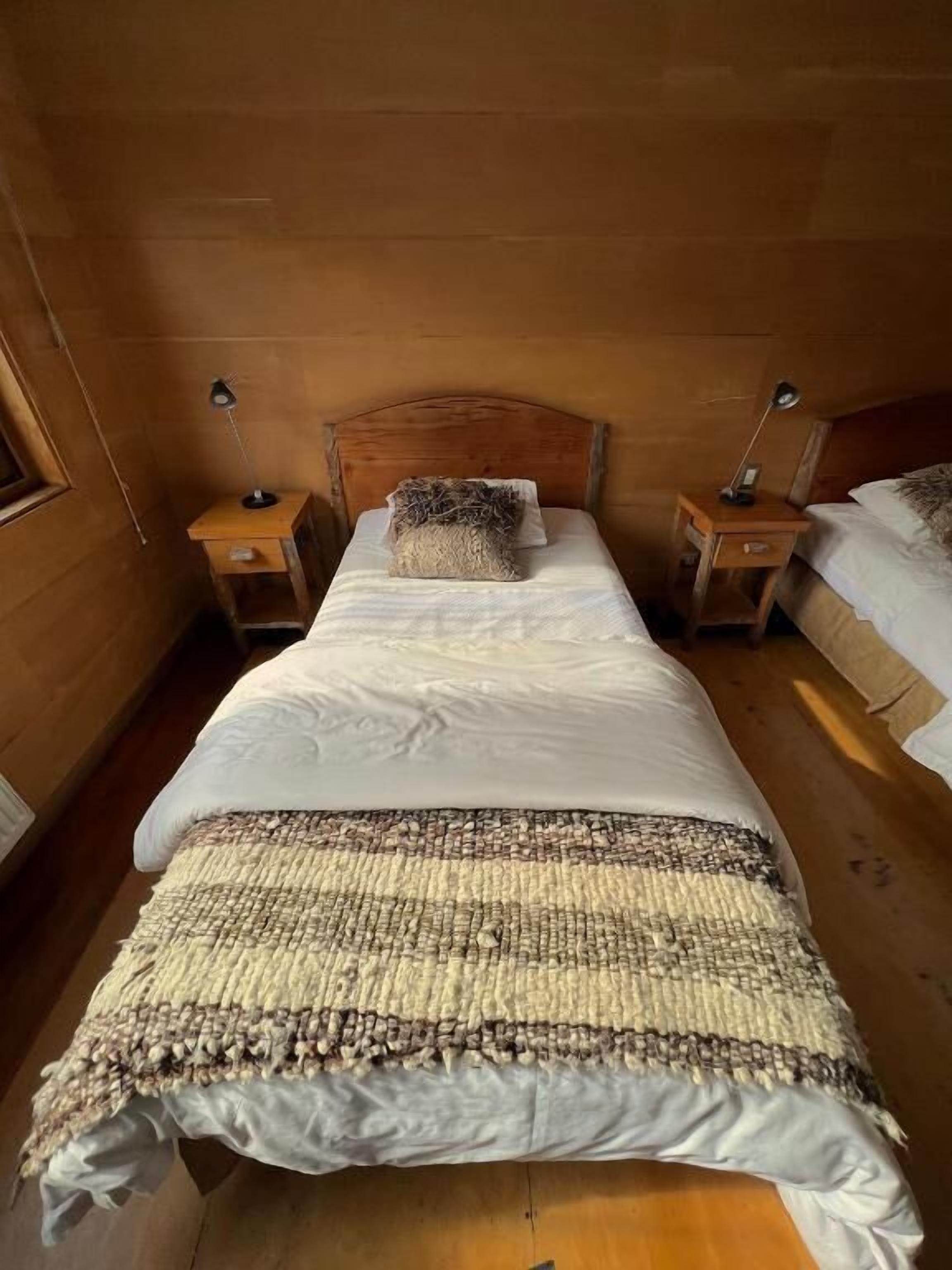 Habitación con cama de cabecera de madera, dos mesitas de noche con lámparas, una manta tejida y suelo de madera.