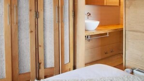 Un dormitorio con cama, un armario de madera con puertas de espejo, una mesita de noche y un baño con lavabo y espejo.