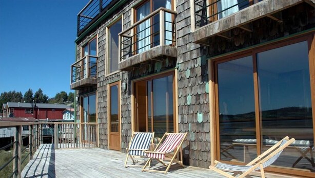 Un edificio de varios pisos con amplias ventanas, una terraza de madera y dos sillones.