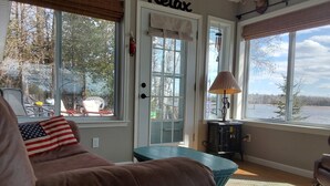 A cozy living room with a brown sofa, a small table, and a lamp. There are large windows with a view of trees and a door with a 'Relax' sign. - Lakefront Home near Alpena Includes Kayaks, Canoe, Rowboat.  Sleeps up to 6 (Alpena)