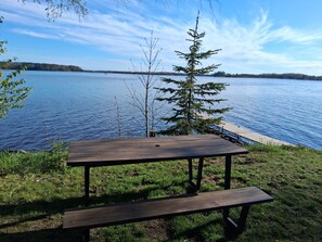 A wooden picnic table with a bench, overlooking a calm lake with a dock and trees in the background.