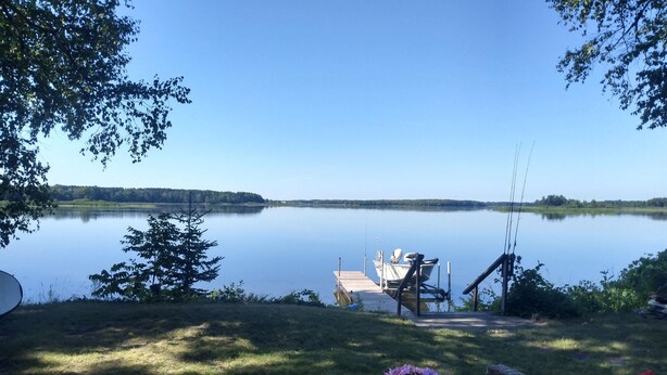 A lakeside view with a dock, a boat, and a wicker chair with a flower arrangement. - Lakefront Home near Alpena Includes Kayaks, Canoe, Rowboat.  Sleeps up to 6 (Alpena)