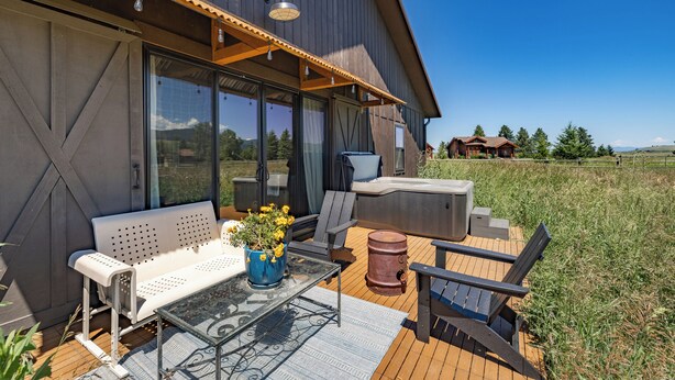 A wooden deck with outdoor furniture, a hot tub, and a large window overlooking a grassy field. - New! Retreat with Hot Tub and Mountain Views near Bozeman! (Gallatin Gateway)