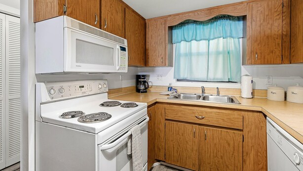 Una cocina compacta con armarios de madera, una estufa blanca, un microondas, un lavabo y una ventana con cortina azul.