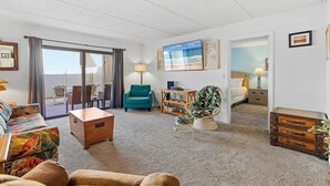 A living room with a sofa, a small wooden table, a teal armchair, a vintage trunk, and a television on the wall. - Oasis 7 (Daytona Beach)