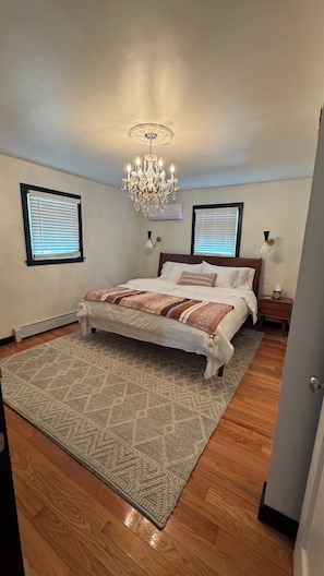 A bedroom with a bed, a chandelier, two wall sconces, a window with blinds, and a radiator.