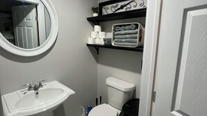 Een badkamer met een witte wastafel, toilet en spiegel. Er zijn planken met handdoeken en toiletartikelen.