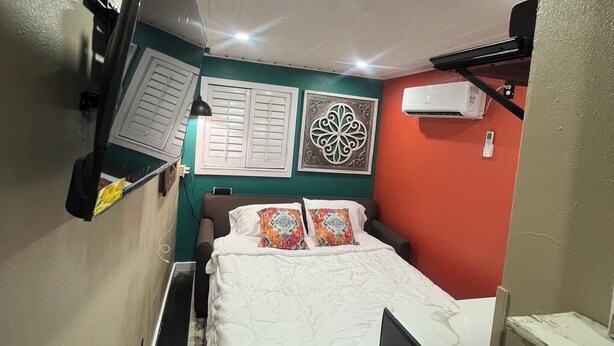 Een compacte hotelkamer met een bed, een flatscreen televisie aan de wand, een raam met luiken en een airconditioner aan de muur.