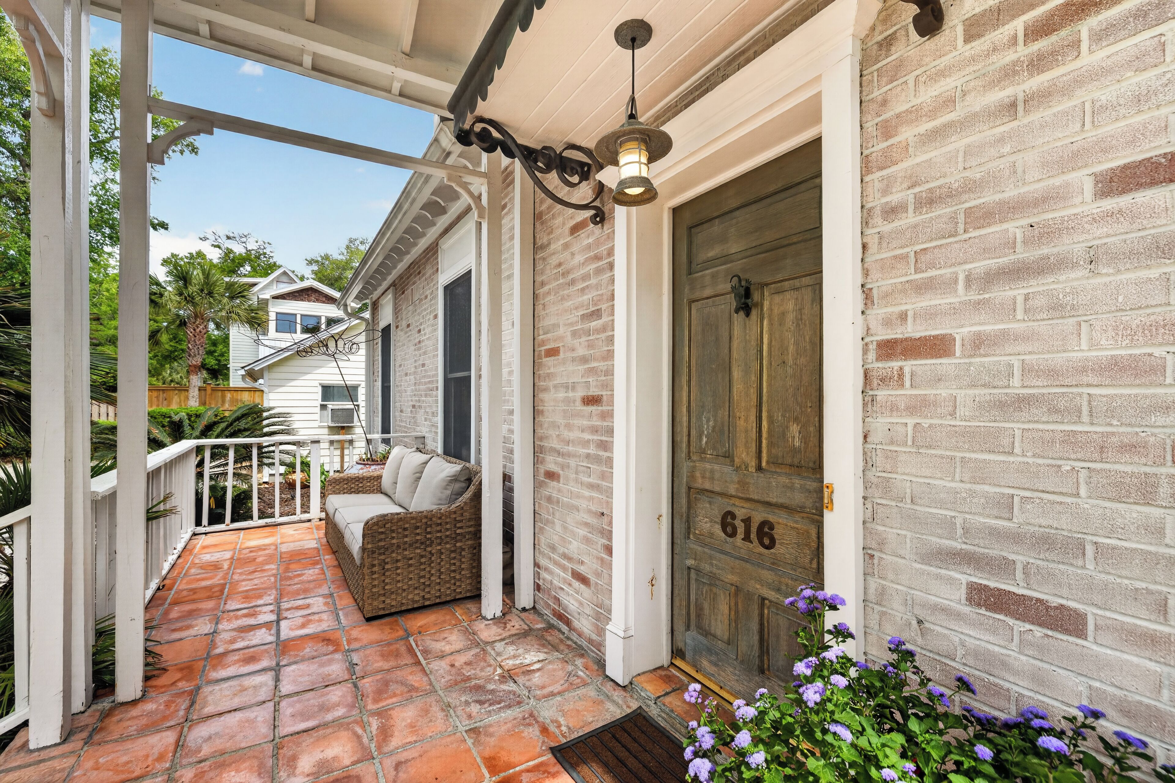 A covered porch with a brick wall, a wooden door with the number 616, and a wicker bench with cushions.