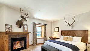 A bedroom with a fireplace, a bed with a wooden headboard, a bench, and deer antlers on the wall. - NEW LISTING | Beautiful Lodge at Mountaineer Square 2 Bedroom (Crested Butte)