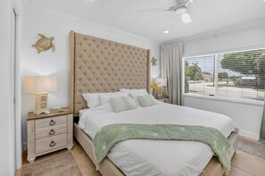A bedroom with a large bed, a nightstand with a lamp, a ceiling fan, and a window with blinds. - BEACH HOME (Boynton Beach)