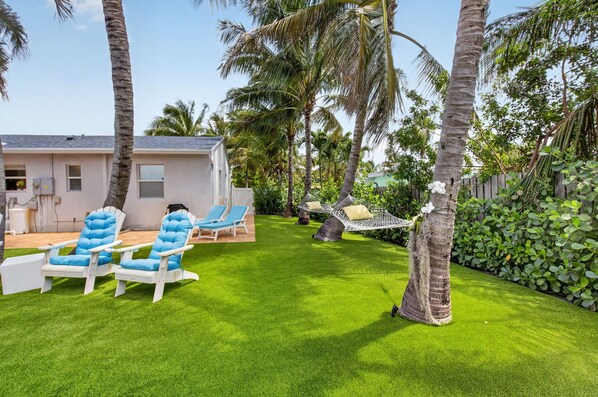 A backyard with artificial grass, two blue lounge chairs, a hammock, and a white building with windows. - BEACH HOME (Boynton Beach)
