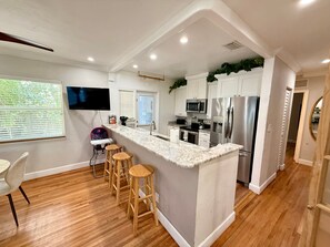 Eine moderne Küche mit Marmorarbeitsplatte, Edelstahlgeräten, Holzbarhocker und einem an der Wand befestigten Fernseher.