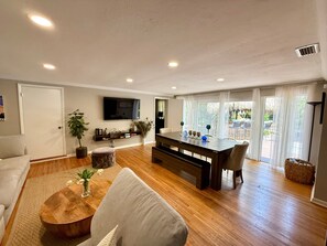 Ein modernes Wohnzimmer mit Holzboden, einer Couch, einem Couchtisch, einem an der Wand befestigten Fernseher und einem Essbereich mit einer Bank und Tisch.