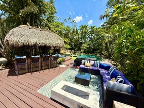 Eine Holzterrasse mit Gartenmöbeln, ein strohgedecktes Gebäude und ein Schwimmbecken, das von üppiger Vegetation umgeben ist.