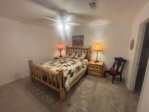 A bedroom with a wooden bed, two bedside tables with lamps, a chair, a ceiling fan, and a painting on the wall.
