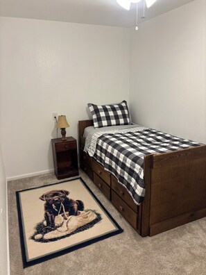 A bedroom with a bed, a nightstand, a lamp, a rug, and a wall socket.