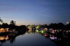 Um canal tranquilo com barcos, palmeiras e edifícios iluminados ao entardecer.
