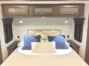 Una cama con cabecera, dos almohadas y una mesita de noche en una habitación con armarios de madera y un ventanal.
