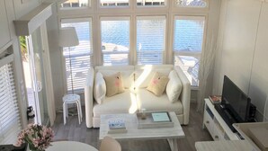 A modern living room with a sofa, coffee table, and TV stand. Large windows offer a view of the water. - Waterfront Tiny Home w/ Pool, Beach, Dock, Kayaks (Key Largo)