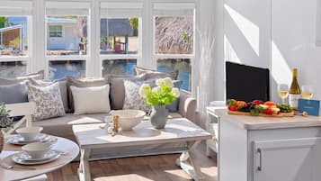 Une pièce baignée de lumière naturelle, avec vue sur un plan d’eau, un coin salon avec des coussins, une table à manger avec un saladier et des couverts, et un comptoir où sont disposés des aliments et une bouteille.
