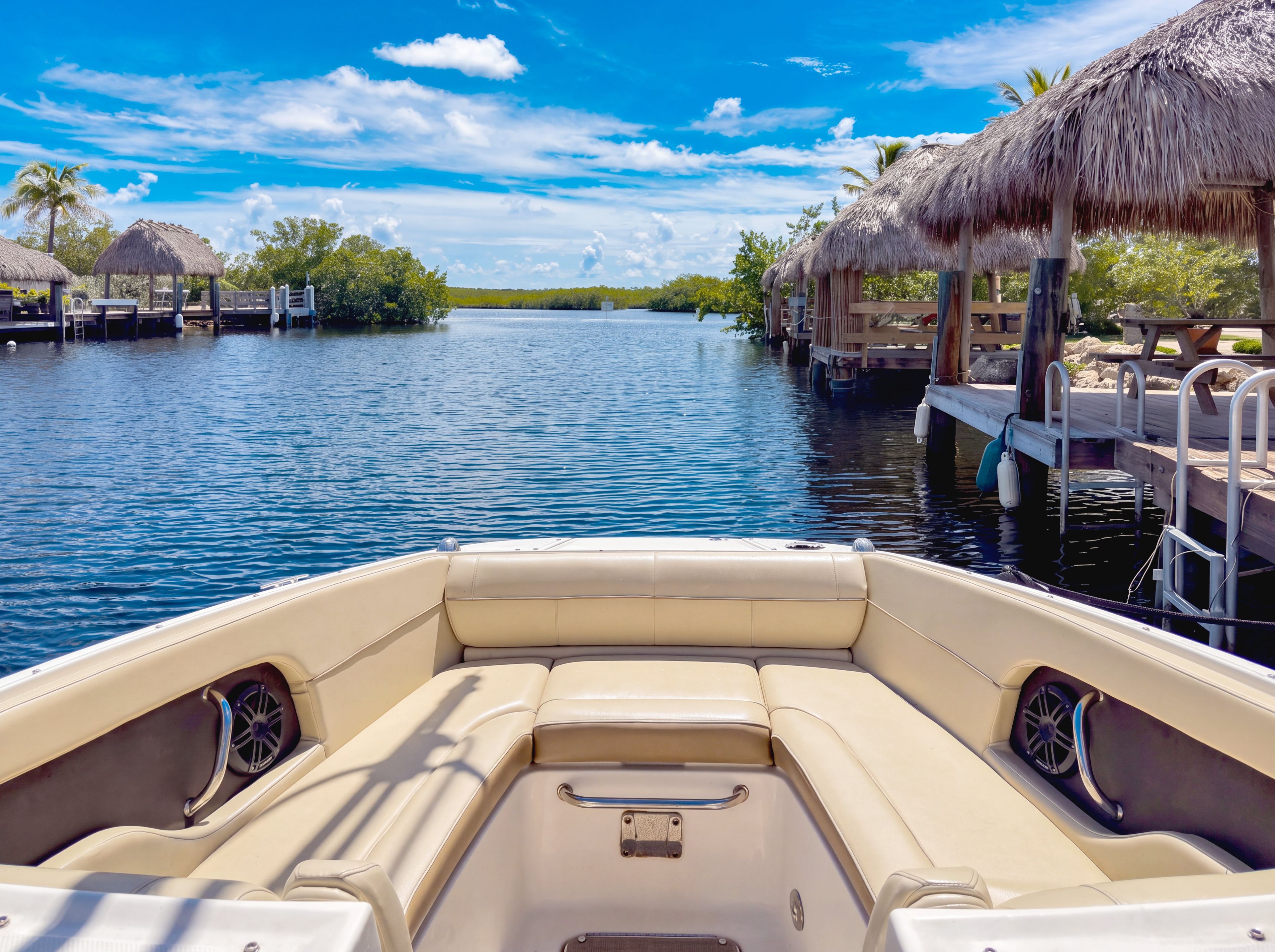 A boat with a beige interior, a thatched-roof structure on the right, and a calm water body with docks.