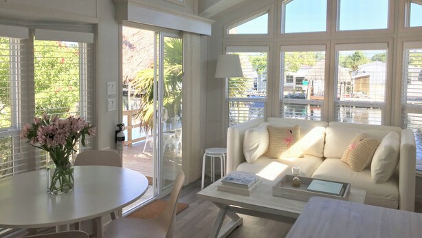 A spacious living room with a large window, a white sofa, a coffee table, and a dining area with a table and chairs. - Waterfront Tiny Home w/ Pool, Beach, Dock, Kayaks (Key Largo)