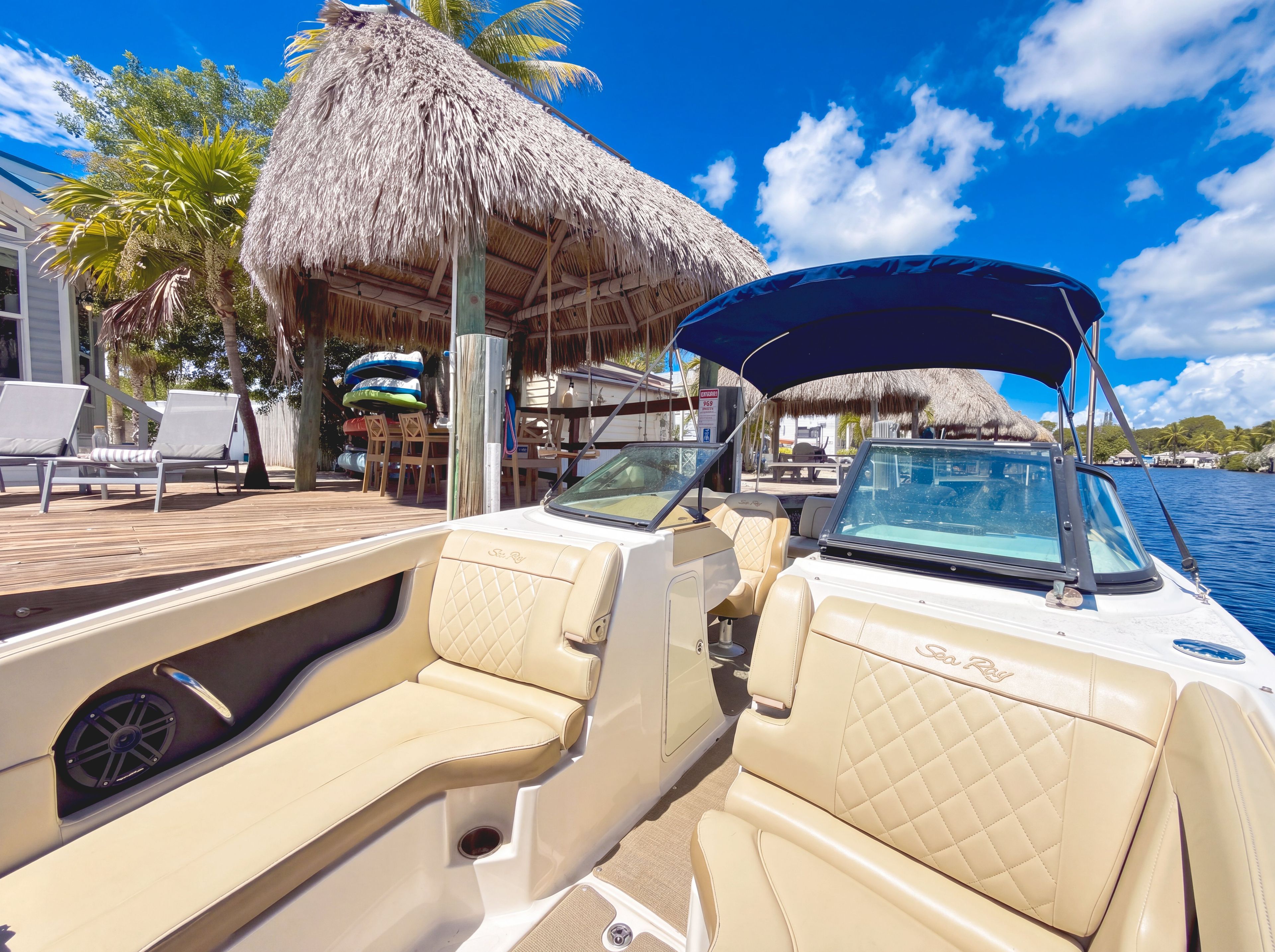 A boat with beige leather seats and a canopy, docked at a resort with a thatched-roof structure and palm trees.