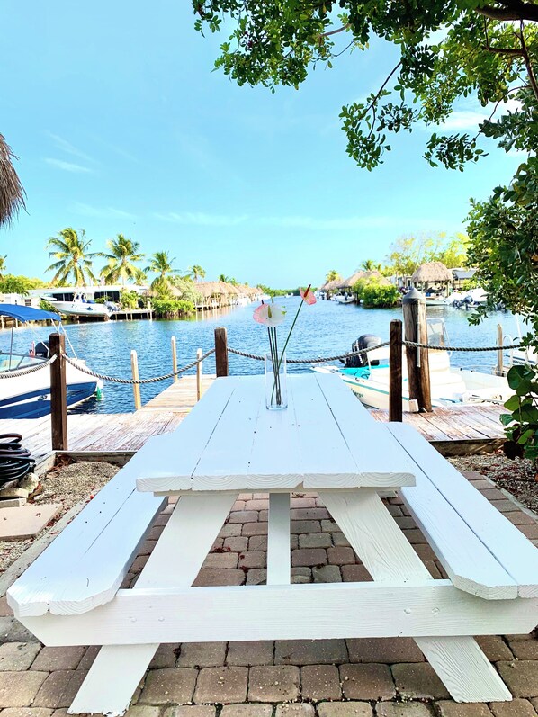 Un muelle de madera con una mesa y sillas, con vista a una masa de agua tranquila con barcos y palmeras.